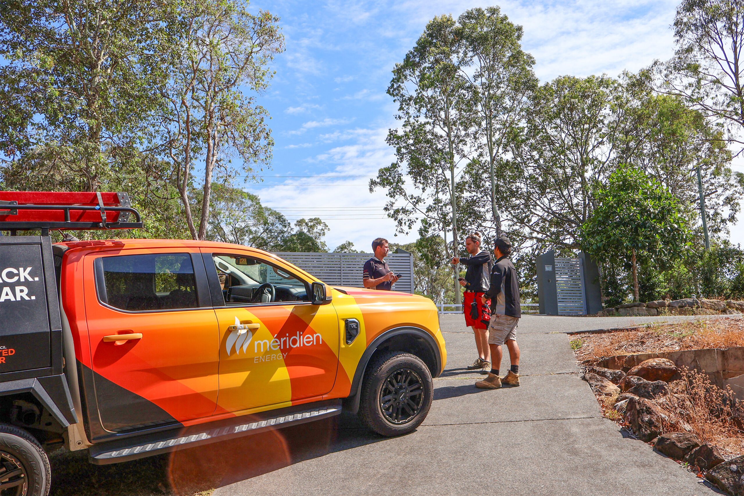 Meridien Energy team, located on the Gold Coast. The solar installers are planning the installation of solar panels and battery system. They are standing next to the bright orange work vehicle, a Ford Ranger ute. They will be working on a Nerang Queensland Home. They will be installing 60x Astronergy N7s 440W Bifacial Solar Panels, 25kW 3-Phase SolaX Hybrid Inverter and two 29.4kWh SolaX Battery stacks. The home is surrounded by luscious gum trees.
